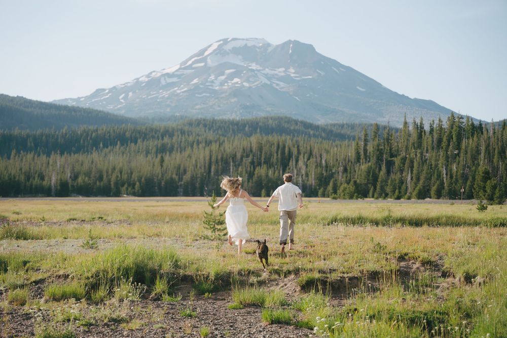 Couple walking hand in hand with their dog across a green meadow with a snow-capped mountain backdrop.