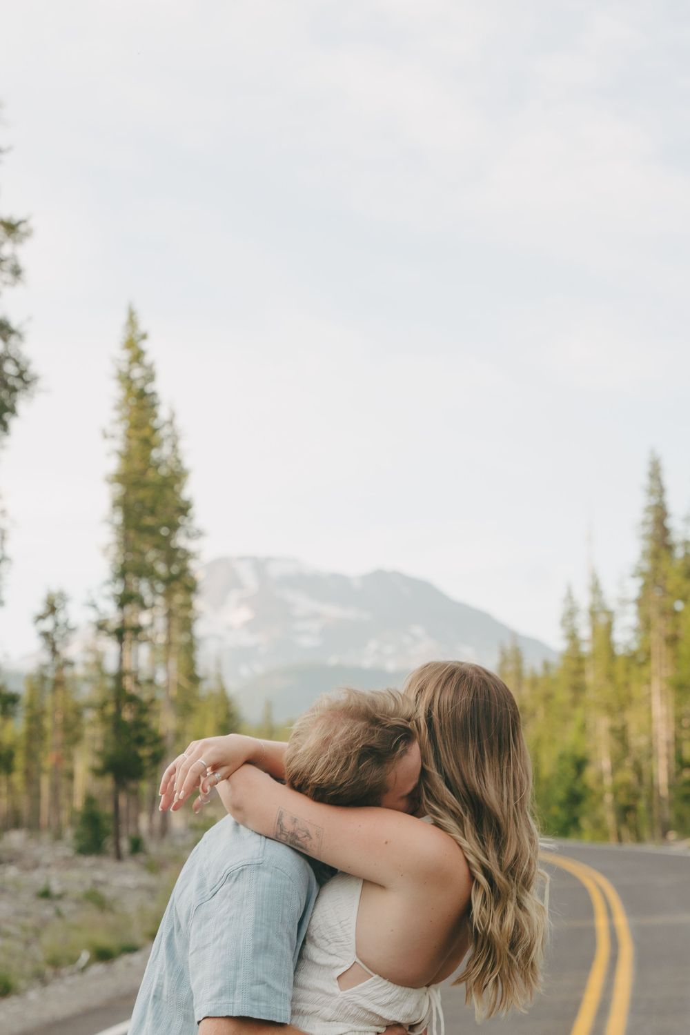 Couple sharing a romantic embrace on a mountain road surrounded by tall pine trees and peaks.