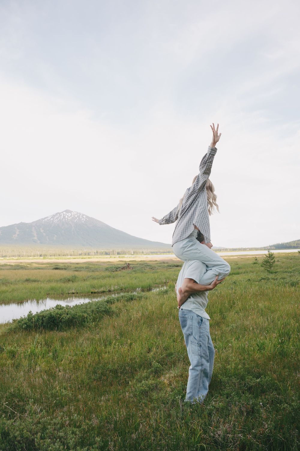 Man lifting woman in a green meadow as she reaches toward the sky with a mountain stream behind them.