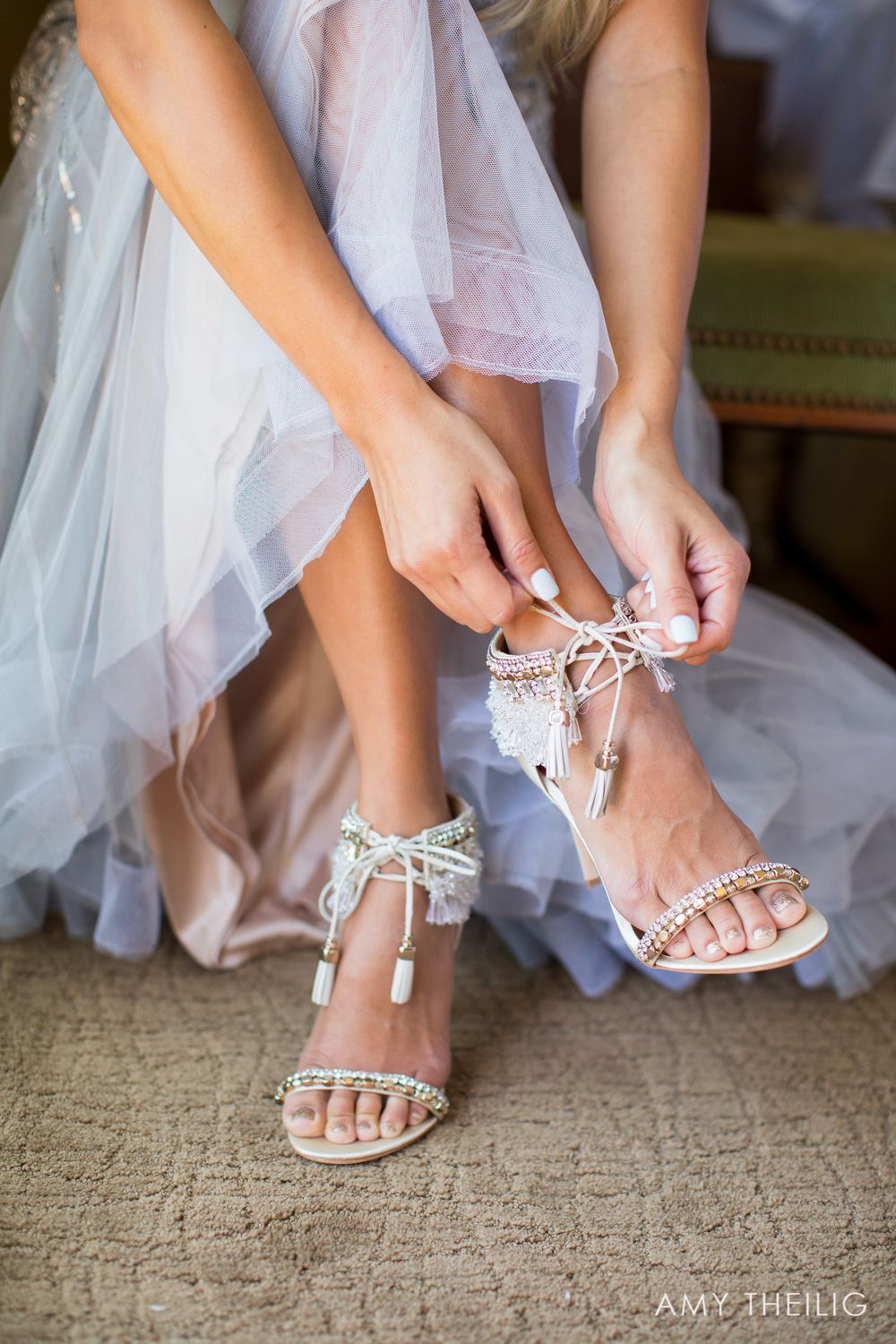 Bride putting on embellished white strappy wedding sandals with tassel details while wearing a tulle gown.
