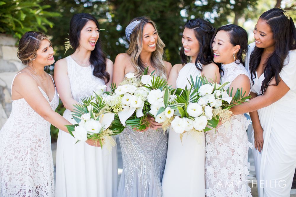 Bride and five bridesmaids in white dresses laughing together while holding white floral bouquets outdoors.