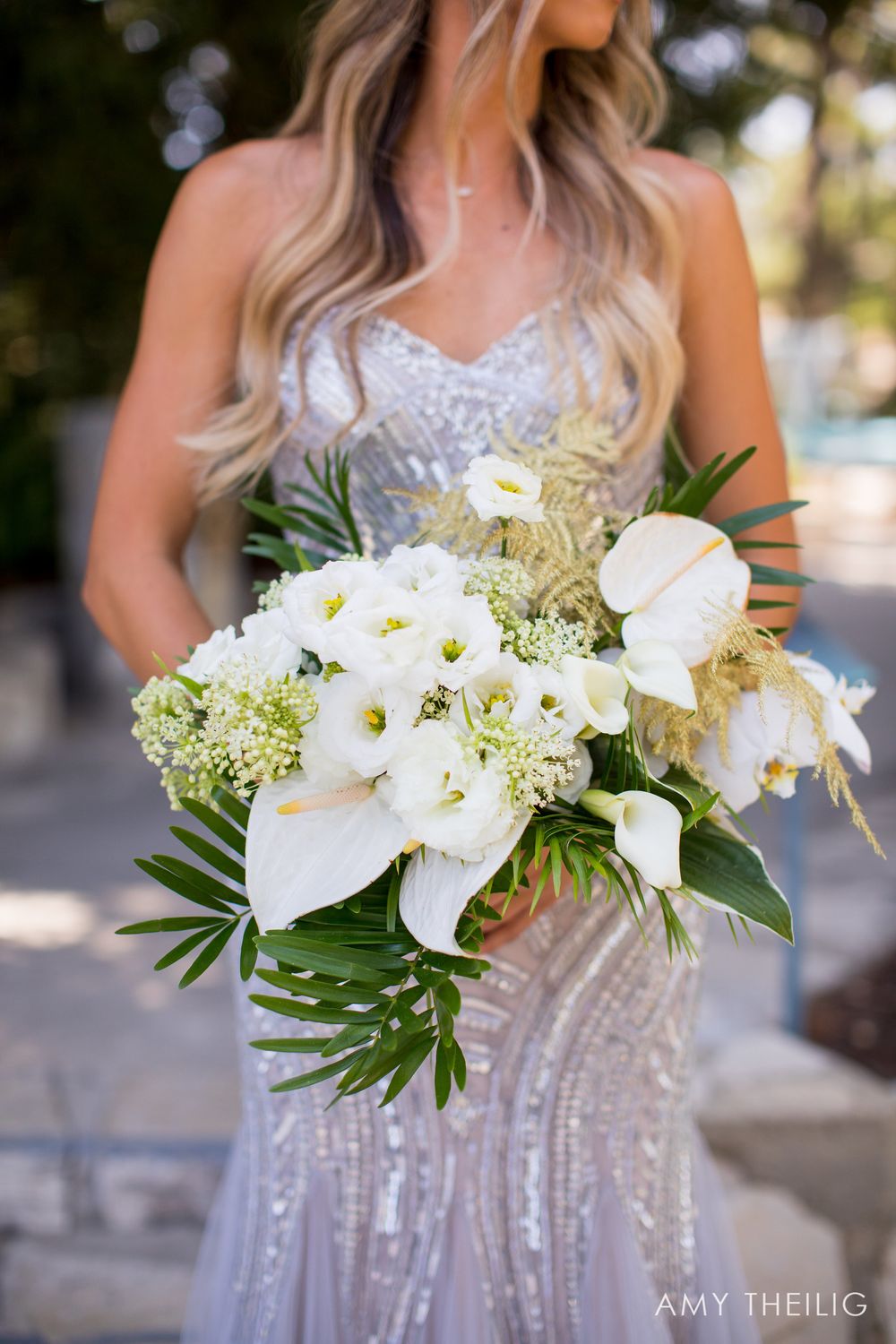 Bride in sequined gown holding lush white tropical bouquet with anthuriums, calla lilies, and palm leaves.