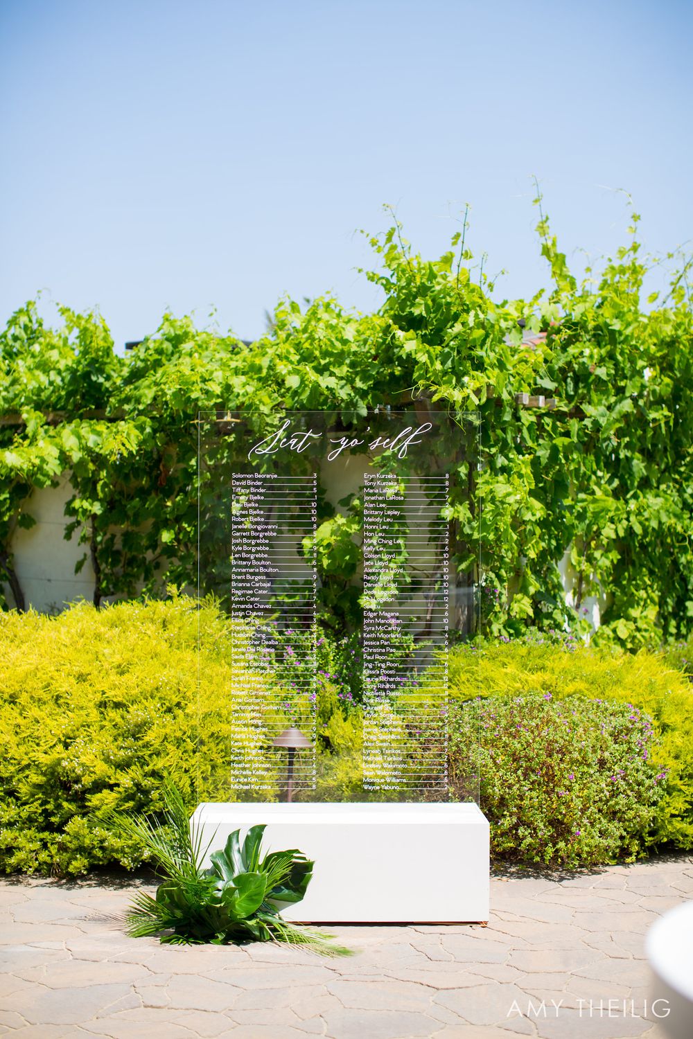 Acrylic wedding seating chart sign with white calligraphy displayed outdoors among lush green hedges.
