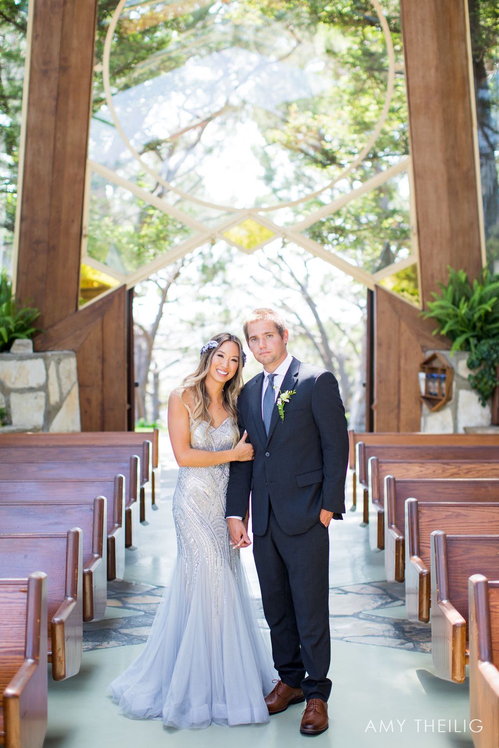Bride in beaded lavender gown and groom in navy suit posing inside a glass chapel with wooden pews.