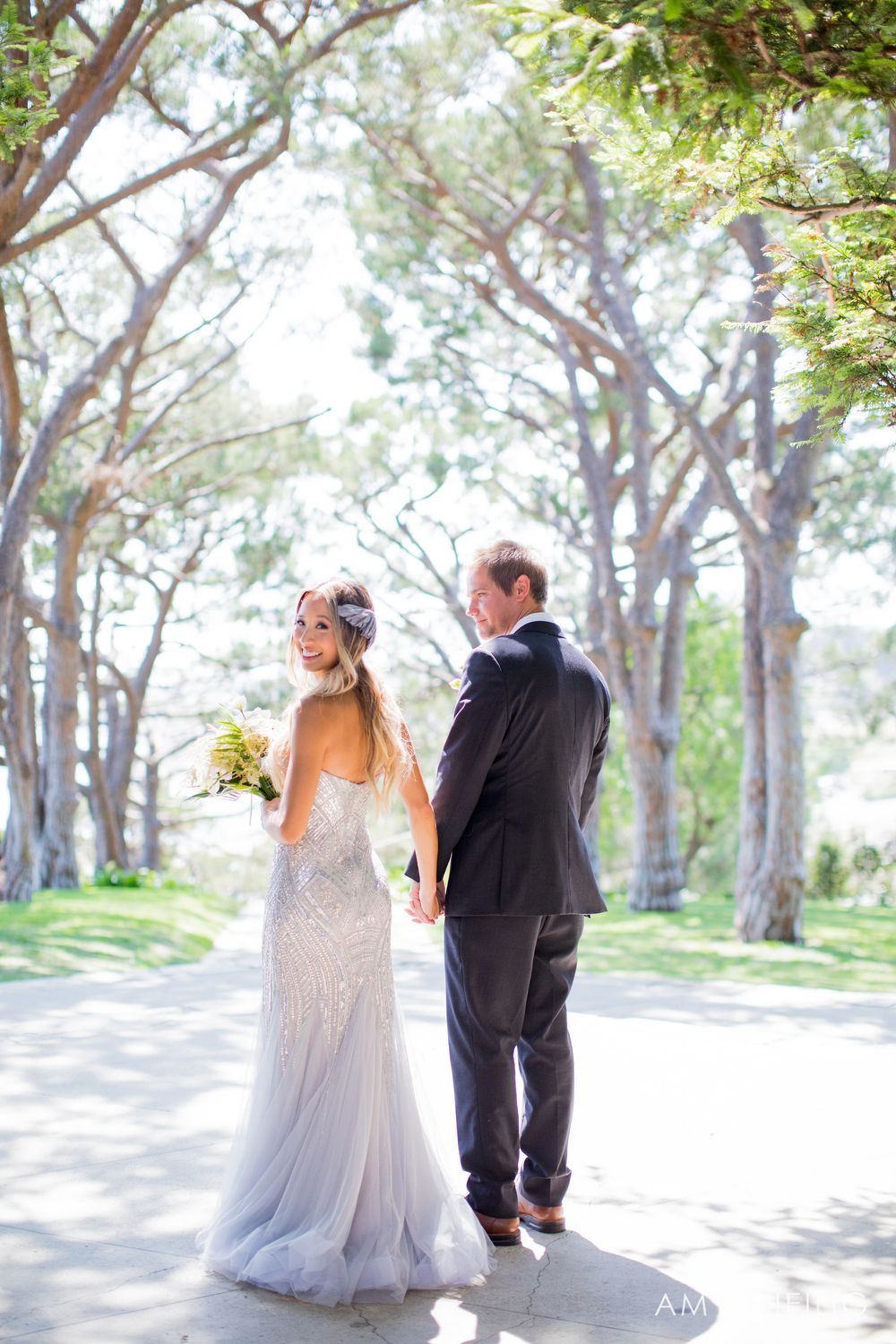 Bride and groom walk hand-in-hand through a sunlit tree-lined path on their wedding day.