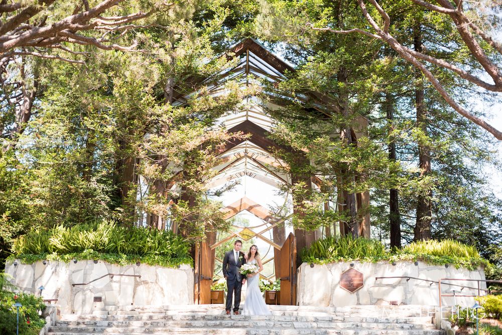 Wedding couple stands before a stunning glass chapel surrounded by tall trees.