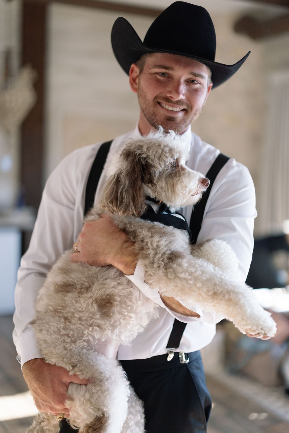 A person wearing a black cowboy hat and white shirt holds and embraces a fluffy white dog.