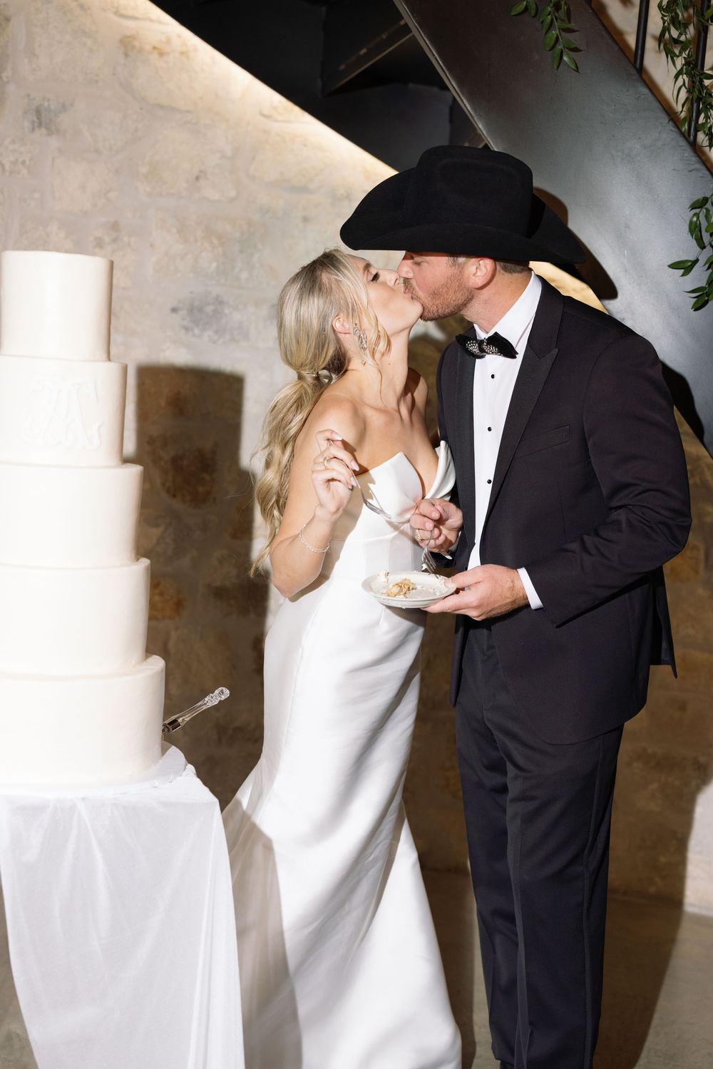 A romantic kissing sequence between a bride in white dress and groom in black tuxedo against a light colored wall.