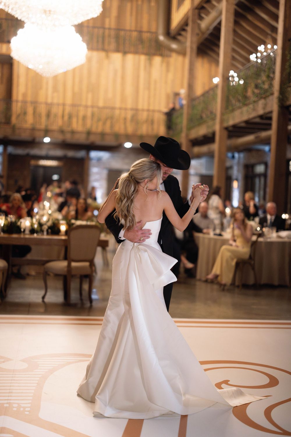 A couple sharing an elegant first dance in a rustic barnhouse venue with a sparkling chandelier overhead.