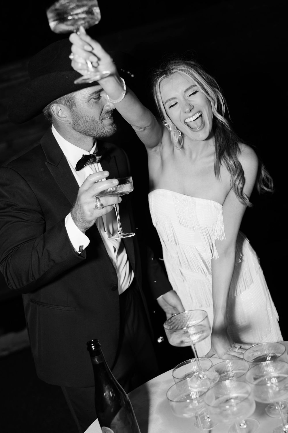 A black and white photo of newlyweds playfully sharing champagne during their wedding celebration.