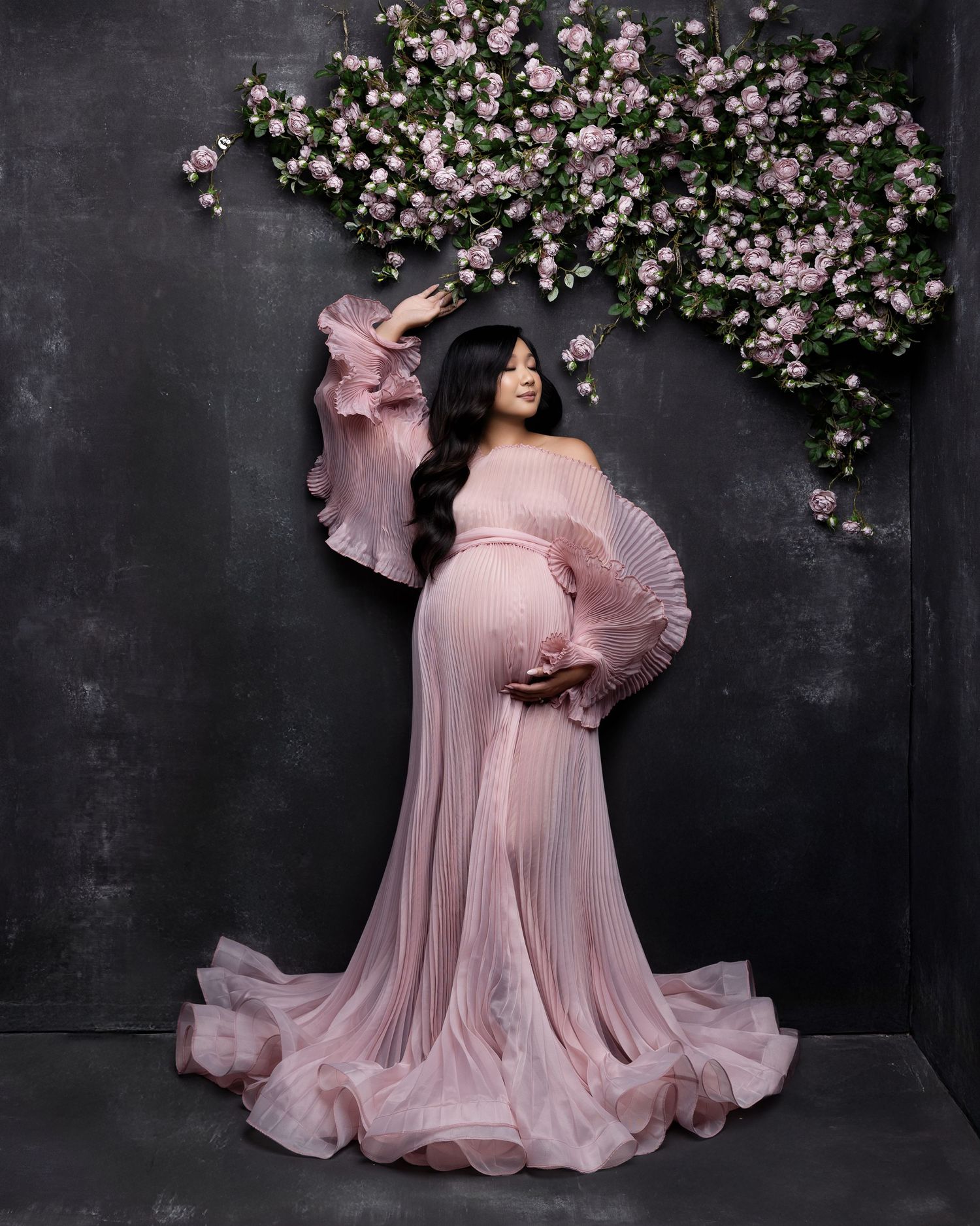 A flowing pink gown with ruffled sleeves stands elegantly beneath a floral arch of pink and white blossoms against a dark wall.