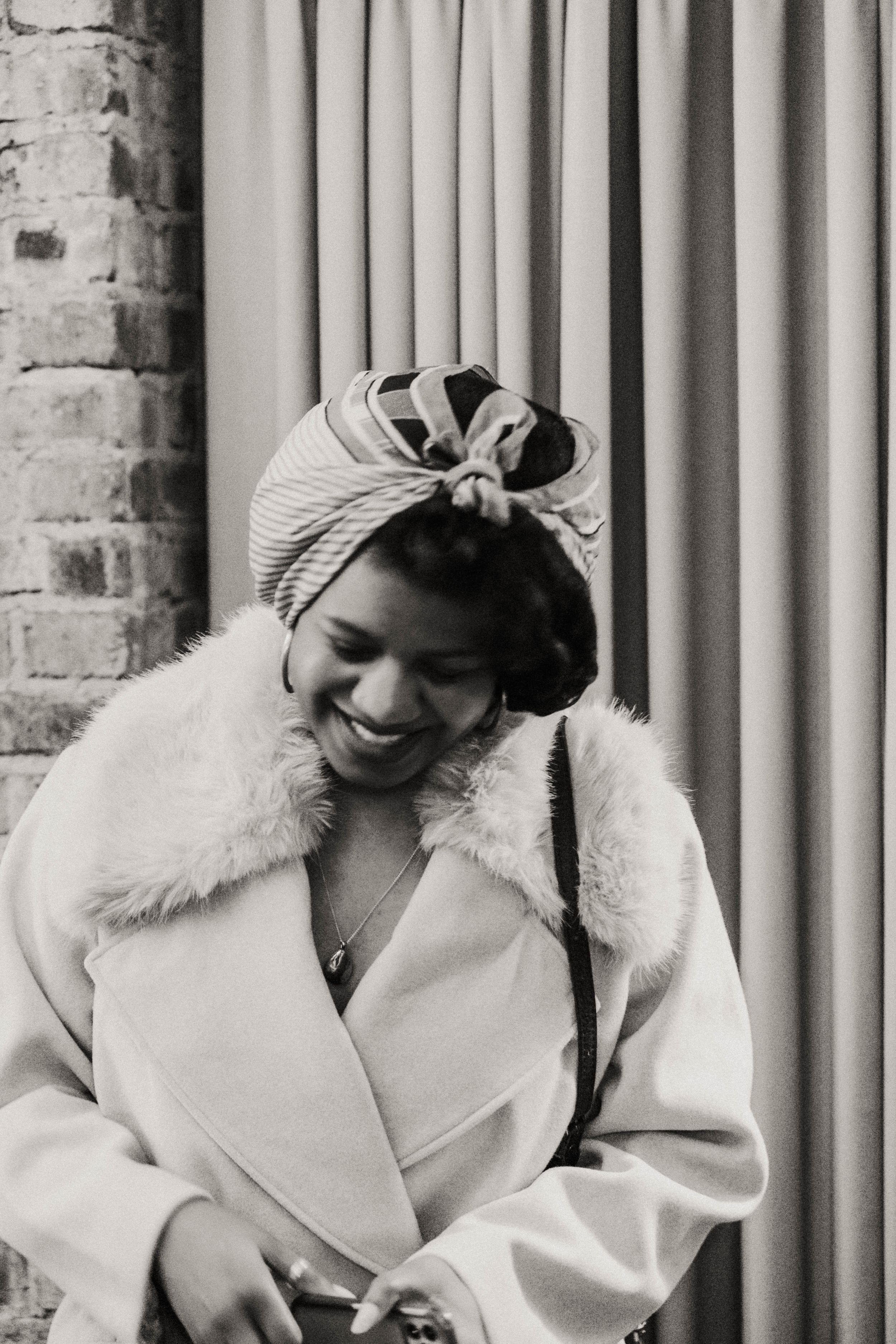Black and white photo of someone in a white fur-collared coat and head wrap smiling while looking down.