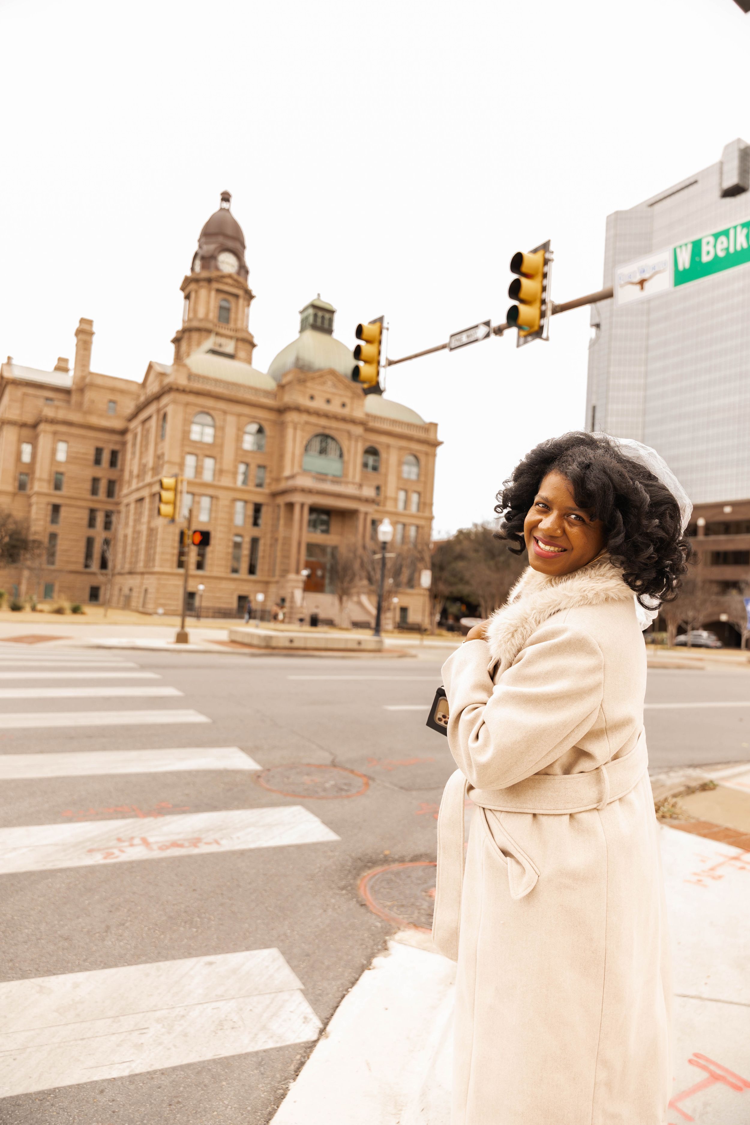 Person in cream winter coat standing at crosswalk with historic courthouse building and traffic lights in background.