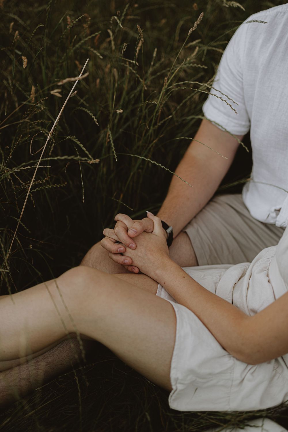 Intimate close-up of intertwined hands resting on legs amid tall grass.