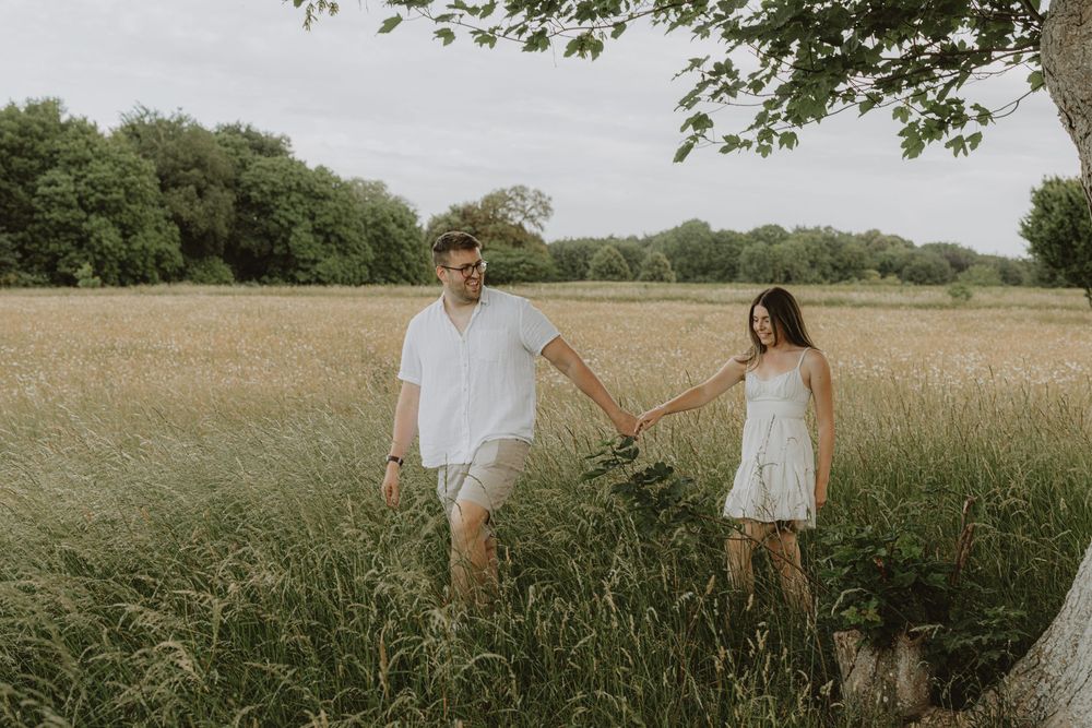 A couple walks hand in hand through a serene meadow filled with wildflowers on an overcast summer day.