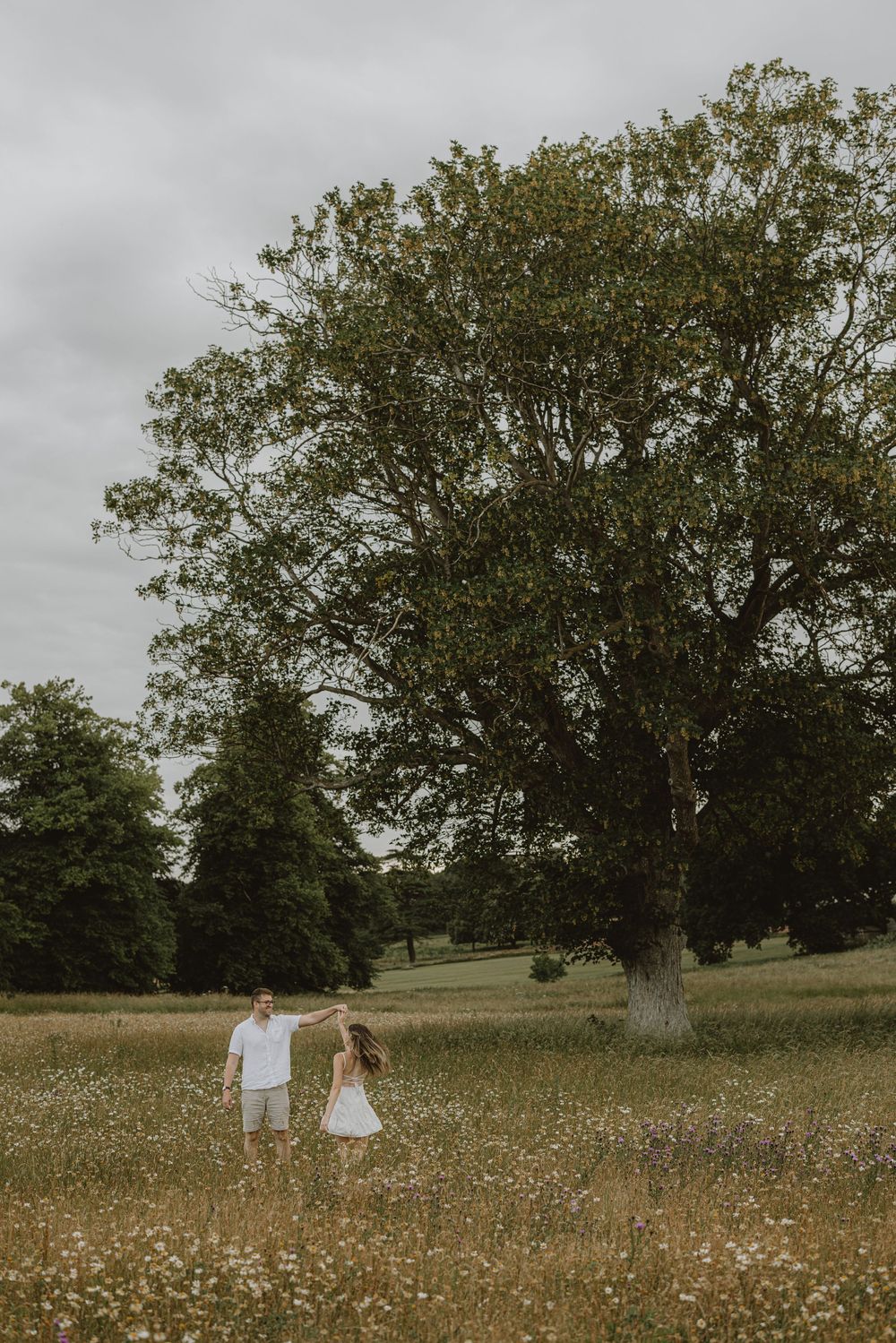 Two figures stroll together through a blooming wildflower field beneath towering trees.