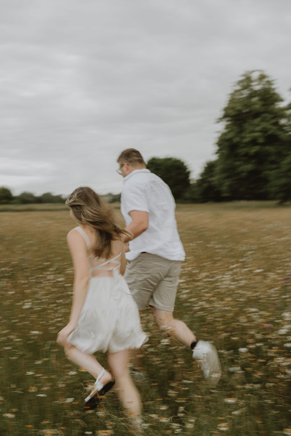 A couple runs playfully through tall grass and wildflowers on a cloudy afternoon.
