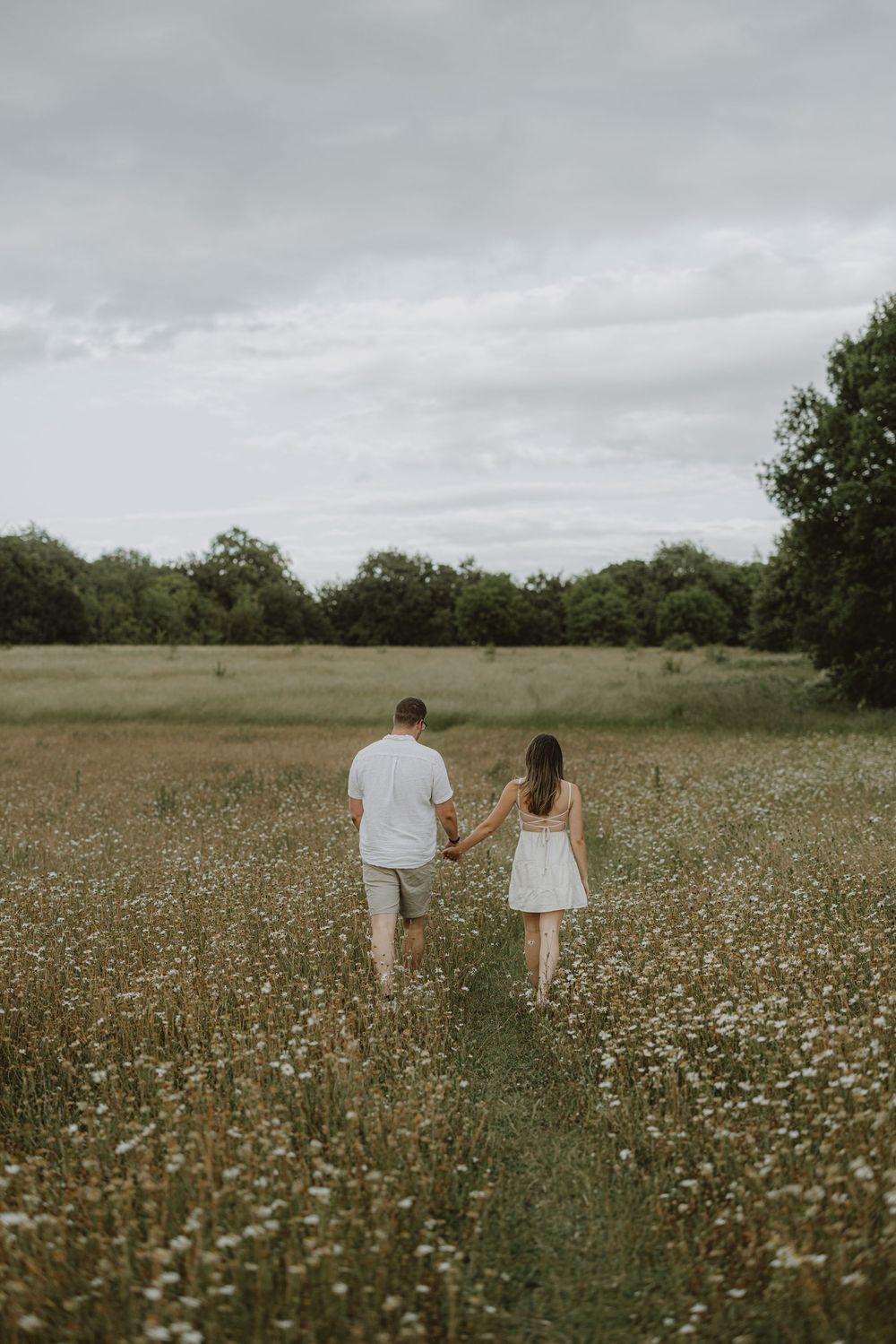 A couple walks through a vast field of daisies under a cloudy summer sky.
