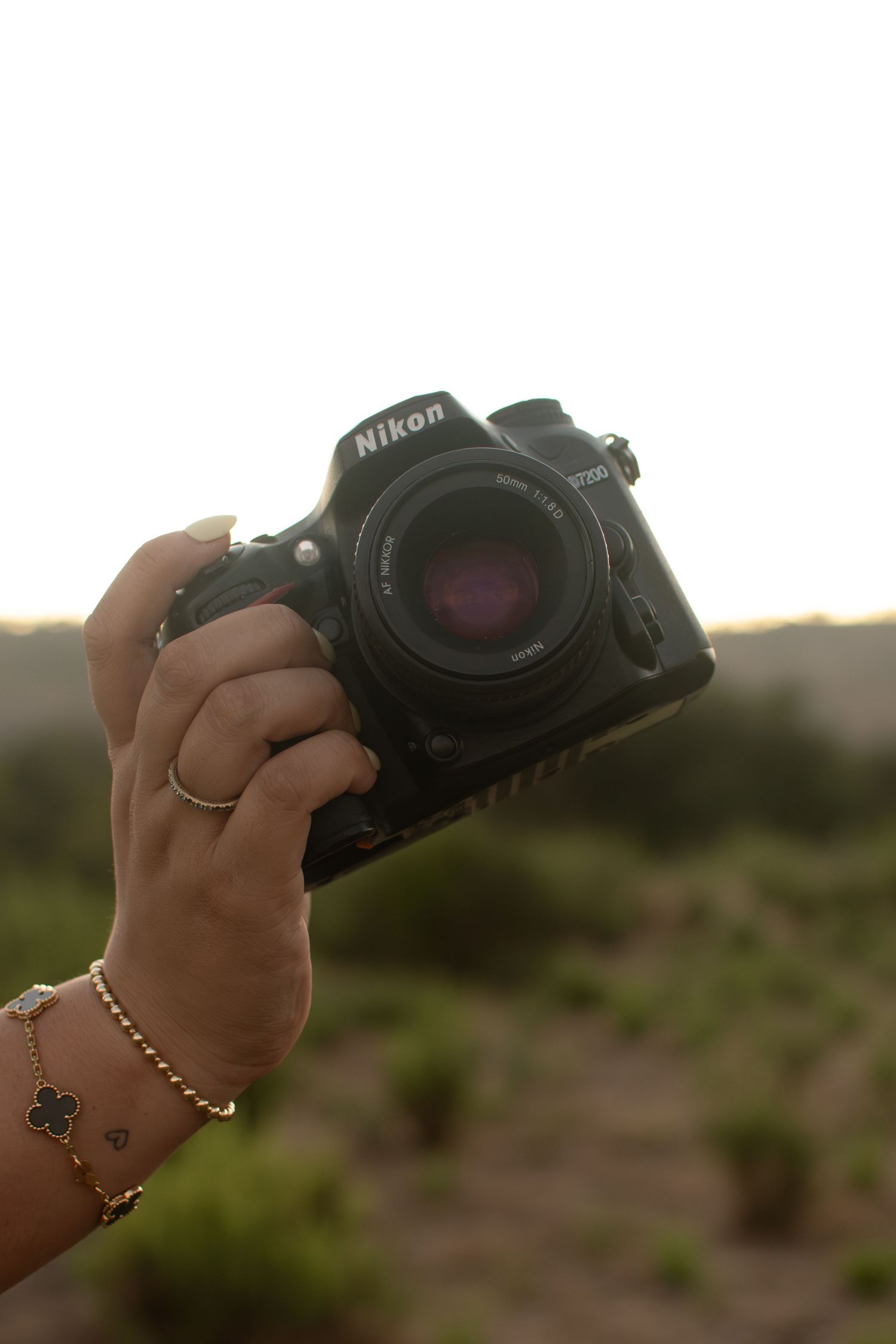 Hand holding a Nikon DSLR camera with gold bracelet and ring against an outdoor landscape.