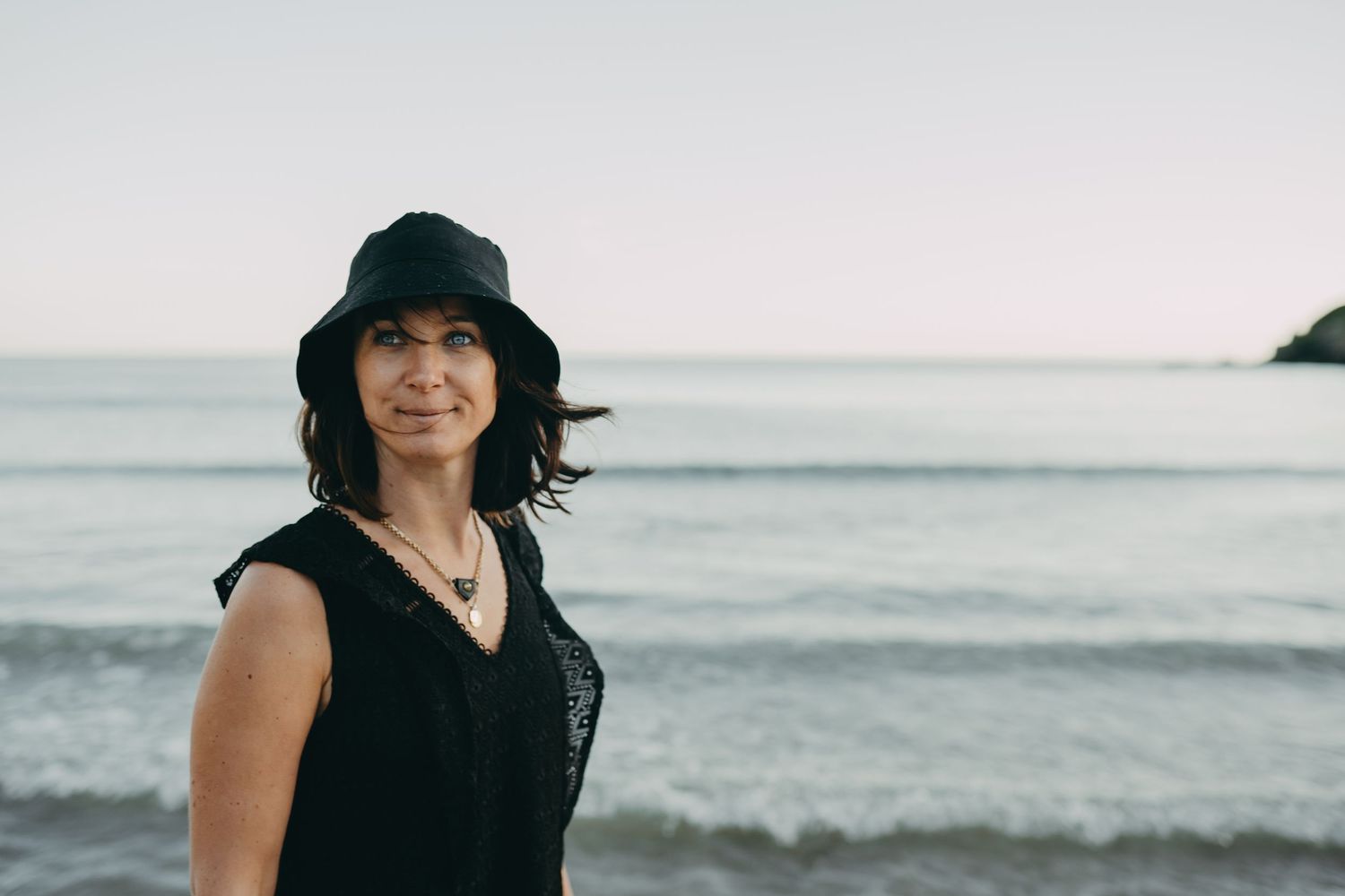 A person in black attire stands against an ocean backdrop at dusk.