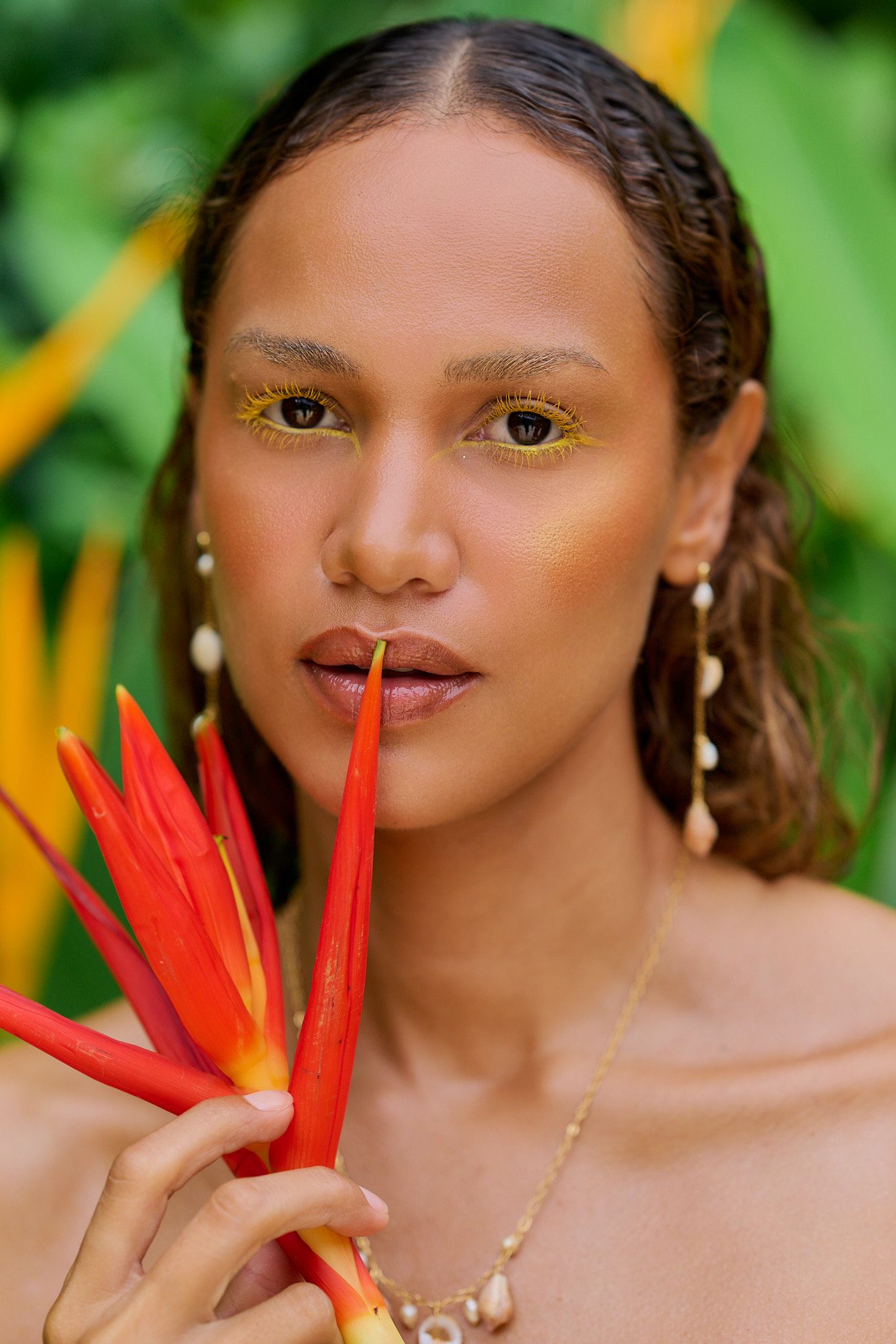 Close-up beauty shot with tropical floral elements, golden eyeshadow makeup and delicate jewelry against green foliage.
