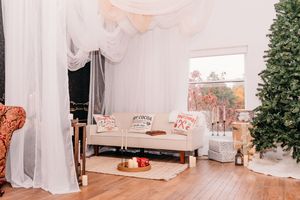 A cozy white living room decorated for Christmas with sheer curtains, a daybed, and a tall Christmas tree in the corner.