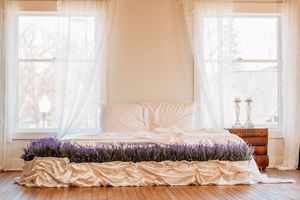 A cozy bedroom with white sheer curtains, a low platform bed with purple bedding, and a wooden nightstand by the window. Photography Studio