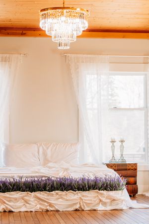 Elegant bedroom with crystal chandelier, white drapes, and wooden floor features lavender accented bedding and candlesticks. Photography