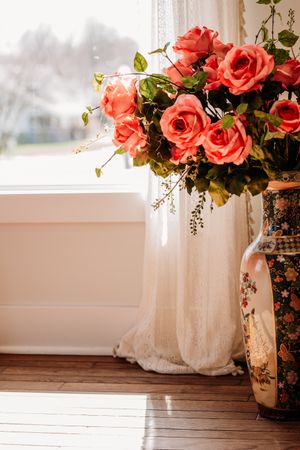 Floral arrangement of coral roses with green foliage in a white vase against a rustic wooden floor and white backdrop. Photography Studio