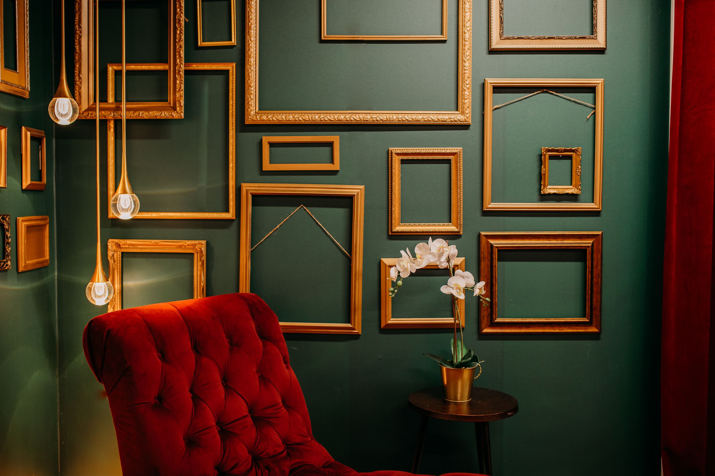 Vintage red velvet armchair against dark green wall with empty gold frames and hanging pendant lights. Photography Studio in Wayland, MI