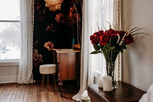 Red roses in glass vase next to window with white curtains and vintage furniture in moody dark interior. Photography Studio in Wayland, MI