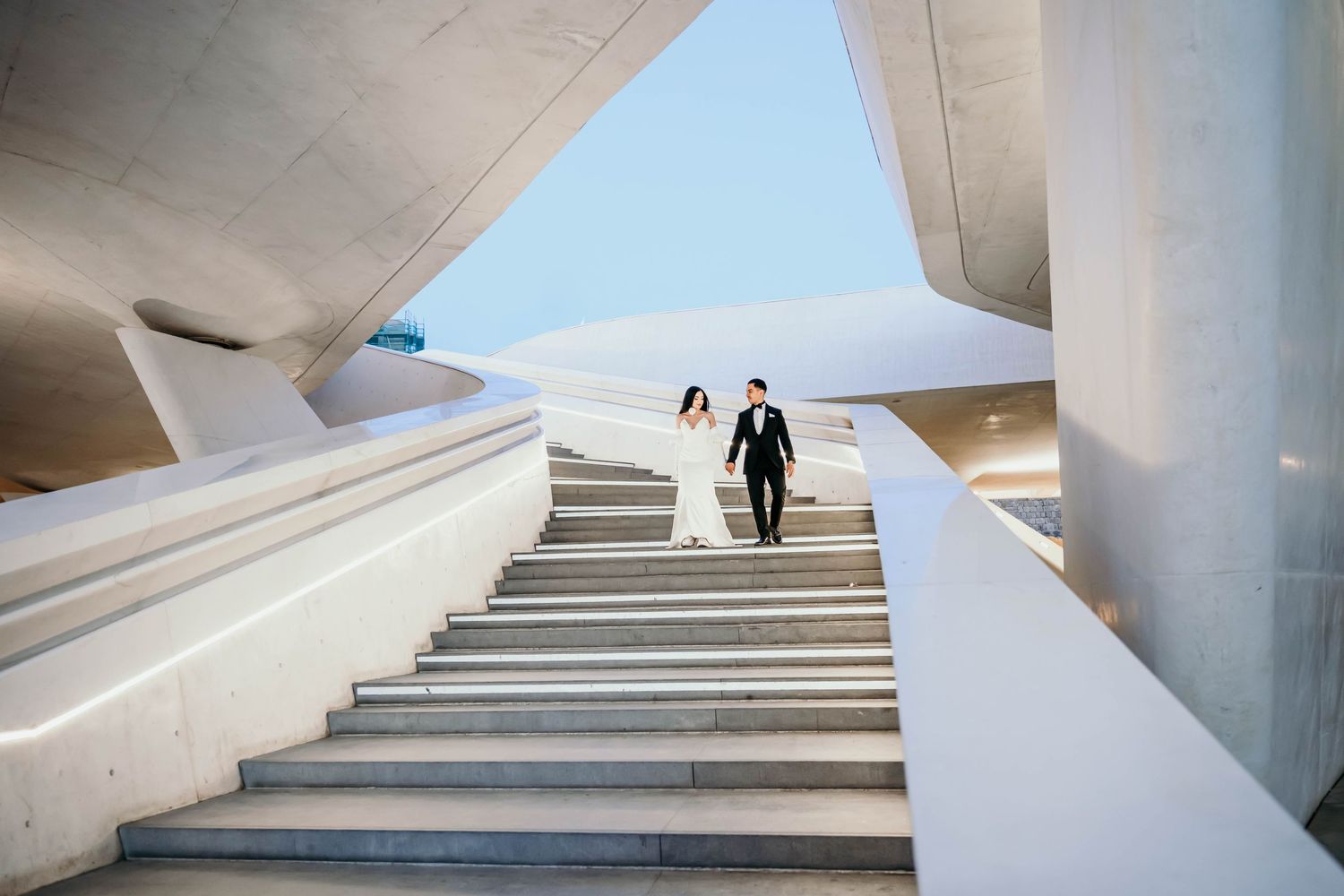 A couple in wedding attire pose on modern white architectural steps with clean geometric lines.