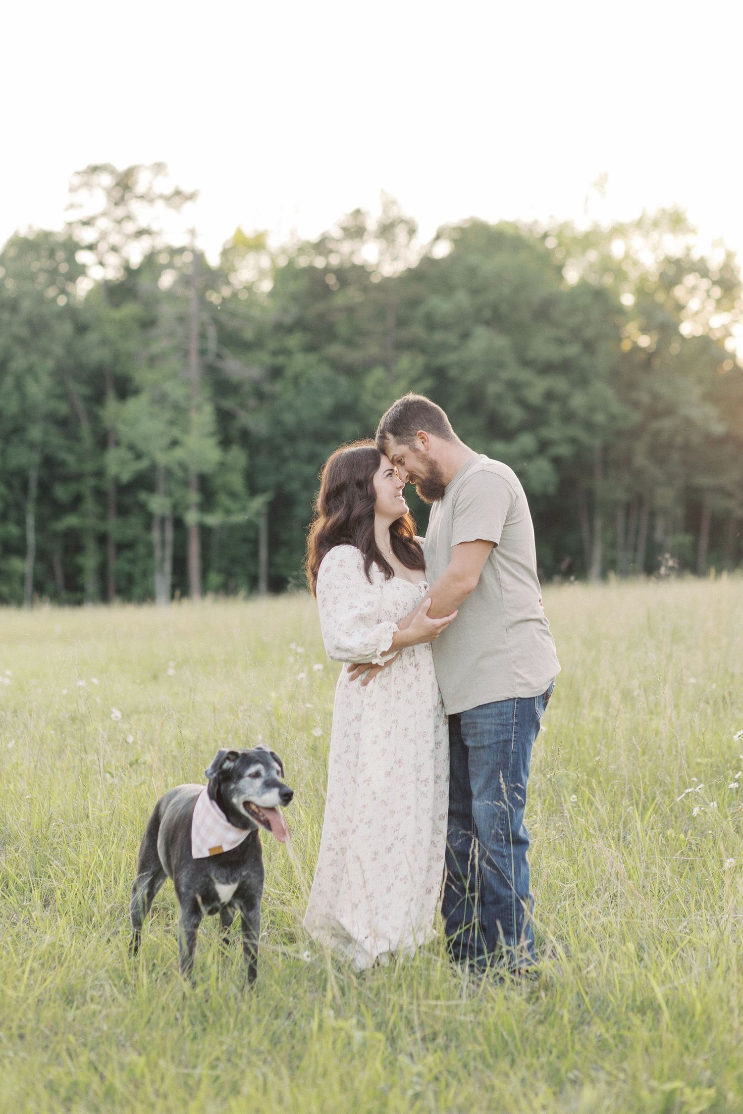 Couple embracing in a sunny meadow with a black dog wearing a bandana beside them.