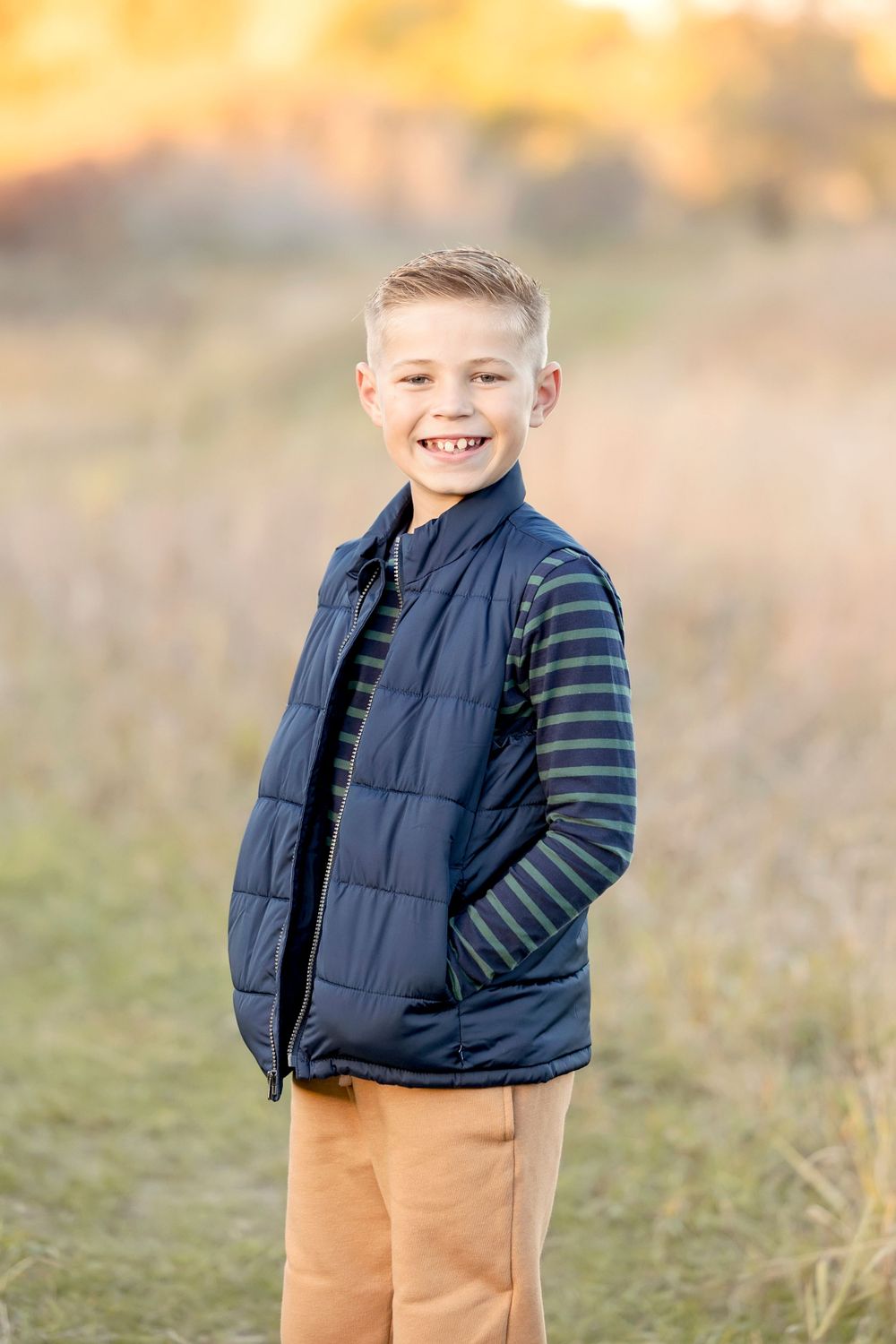 Young child wearing a navy vest and striped shirt poses outdoors in golden autumn light. Clear Lake Photographer. Mason City Photographer.