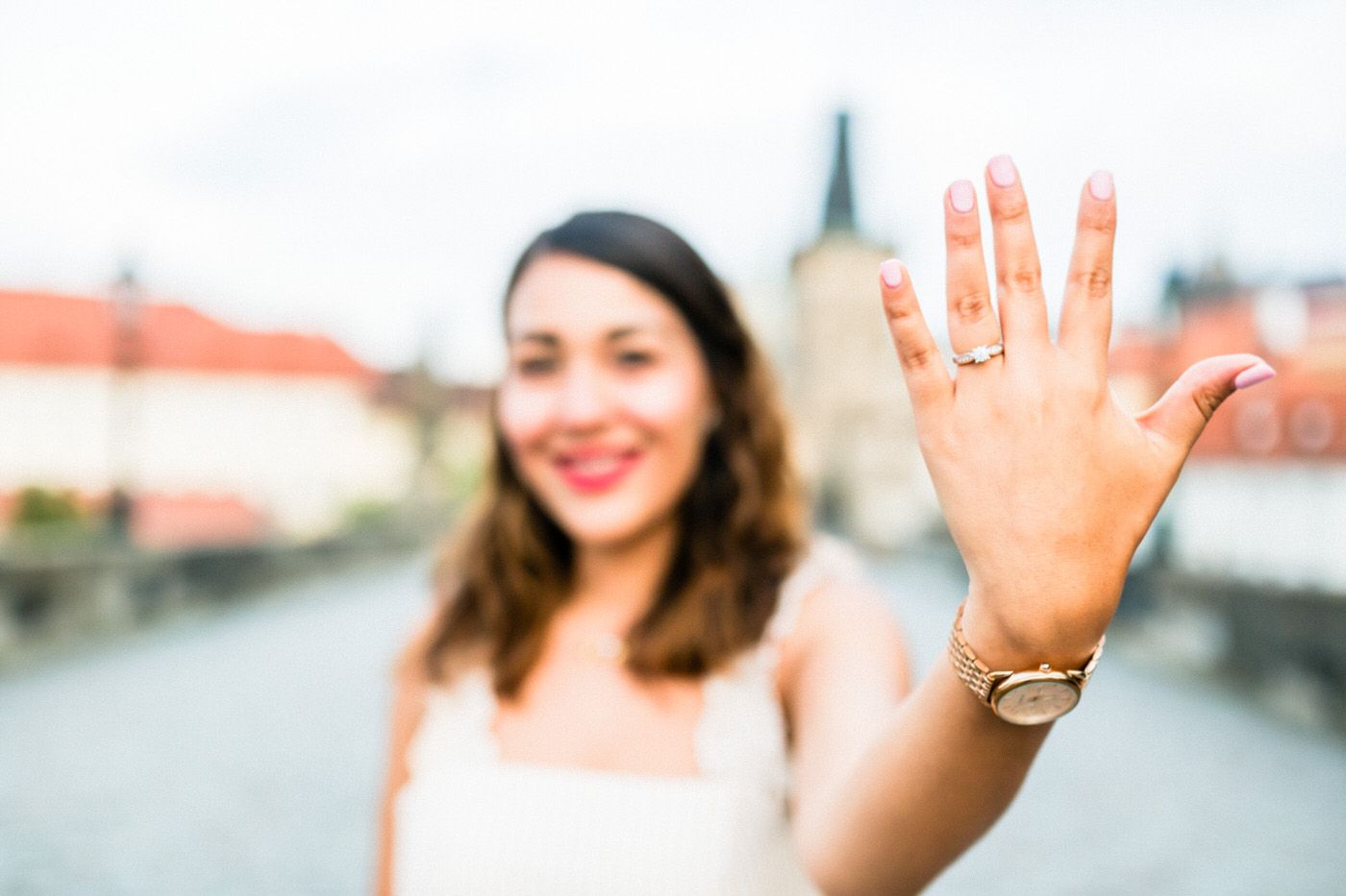 Close up of a hand showing off an engagement ring with a blurred cityscape in the background.