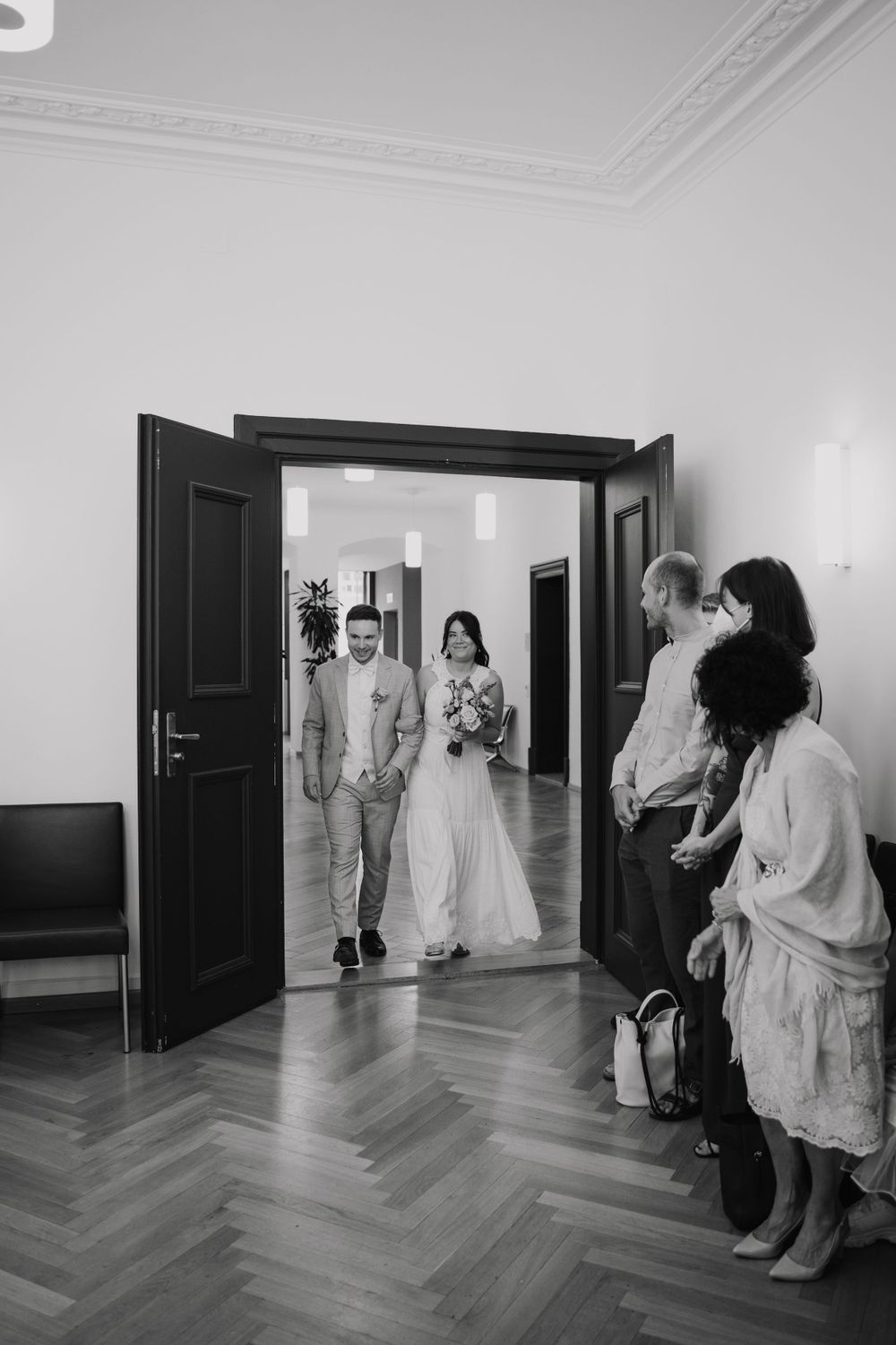 Black and white photo of a bride and groom entering a room greeted by guests at a wedding ceremony. Berlin
