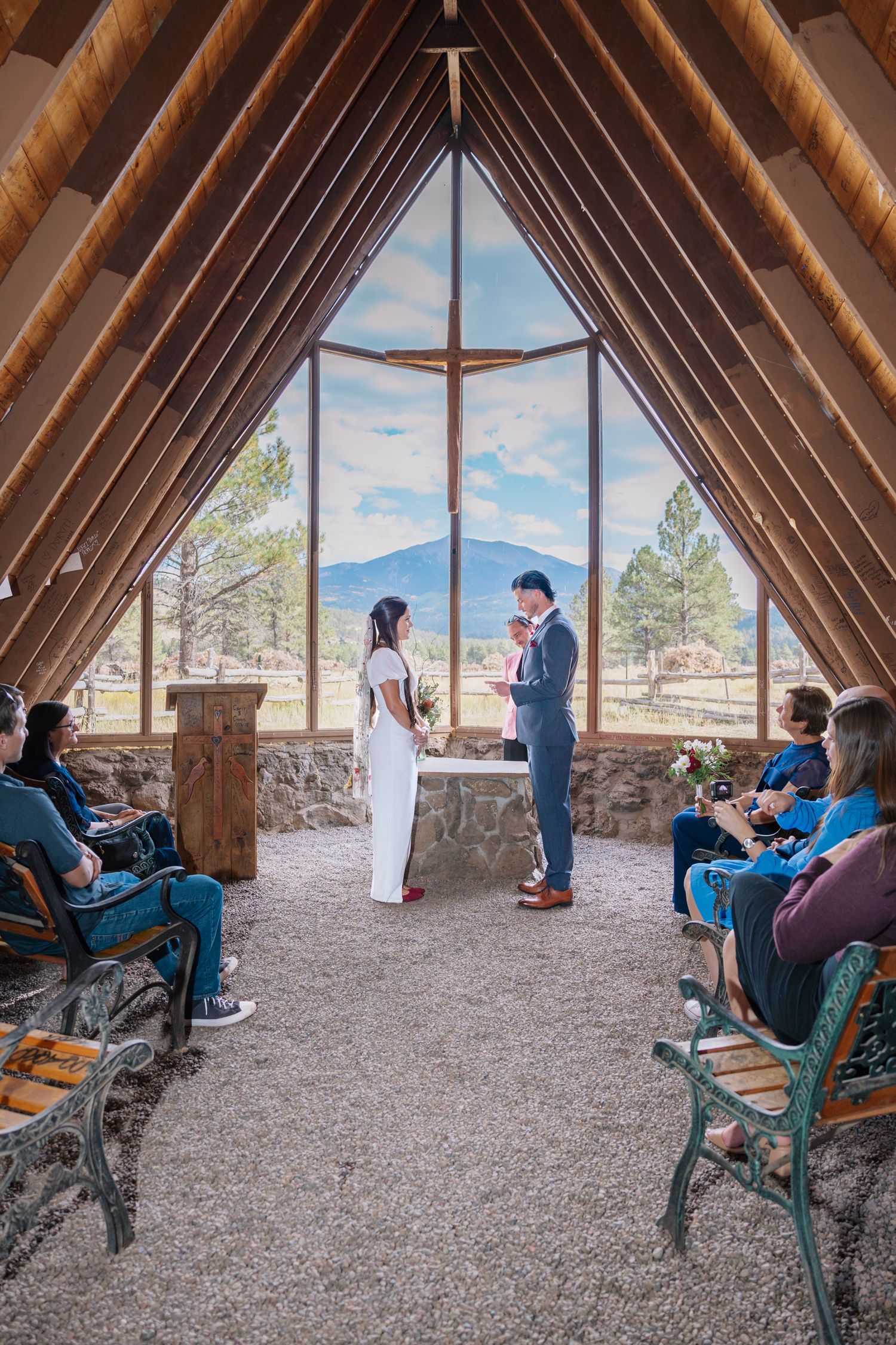 Wedding ceremony inside rustic chapel with dramatic triangular window frame overlooking mountain landscape.