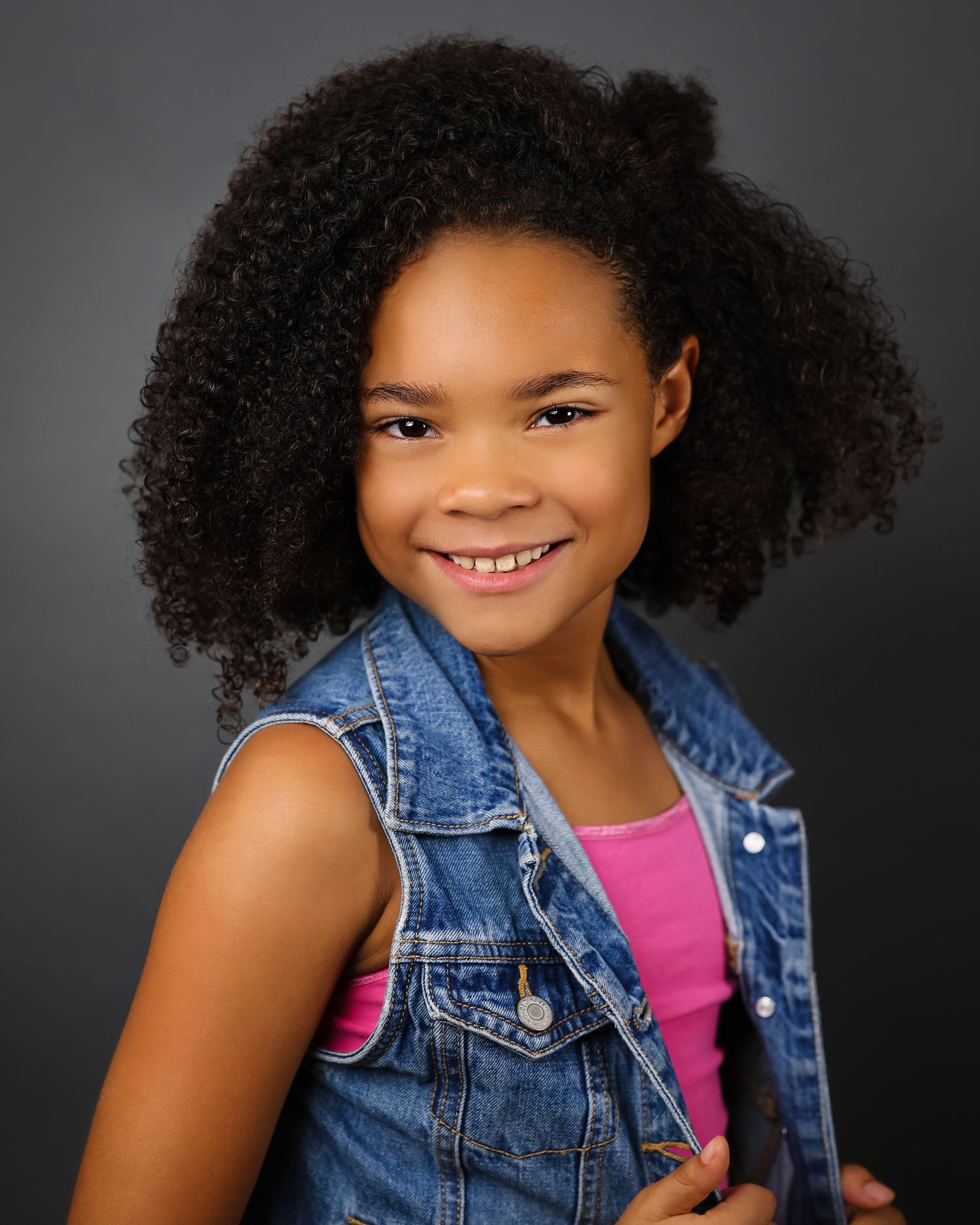 Portrait of a young actress wearing denim vest and pink top against dark background.