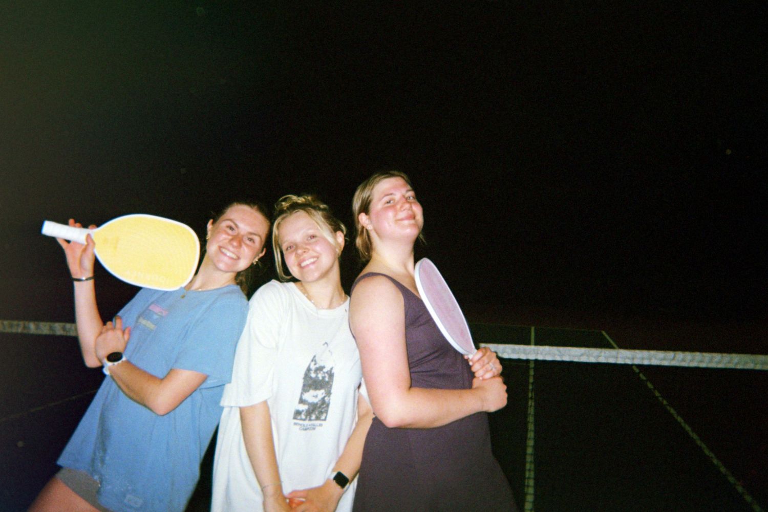 Three friends posing together at night while holding a yellow paddle near a net.