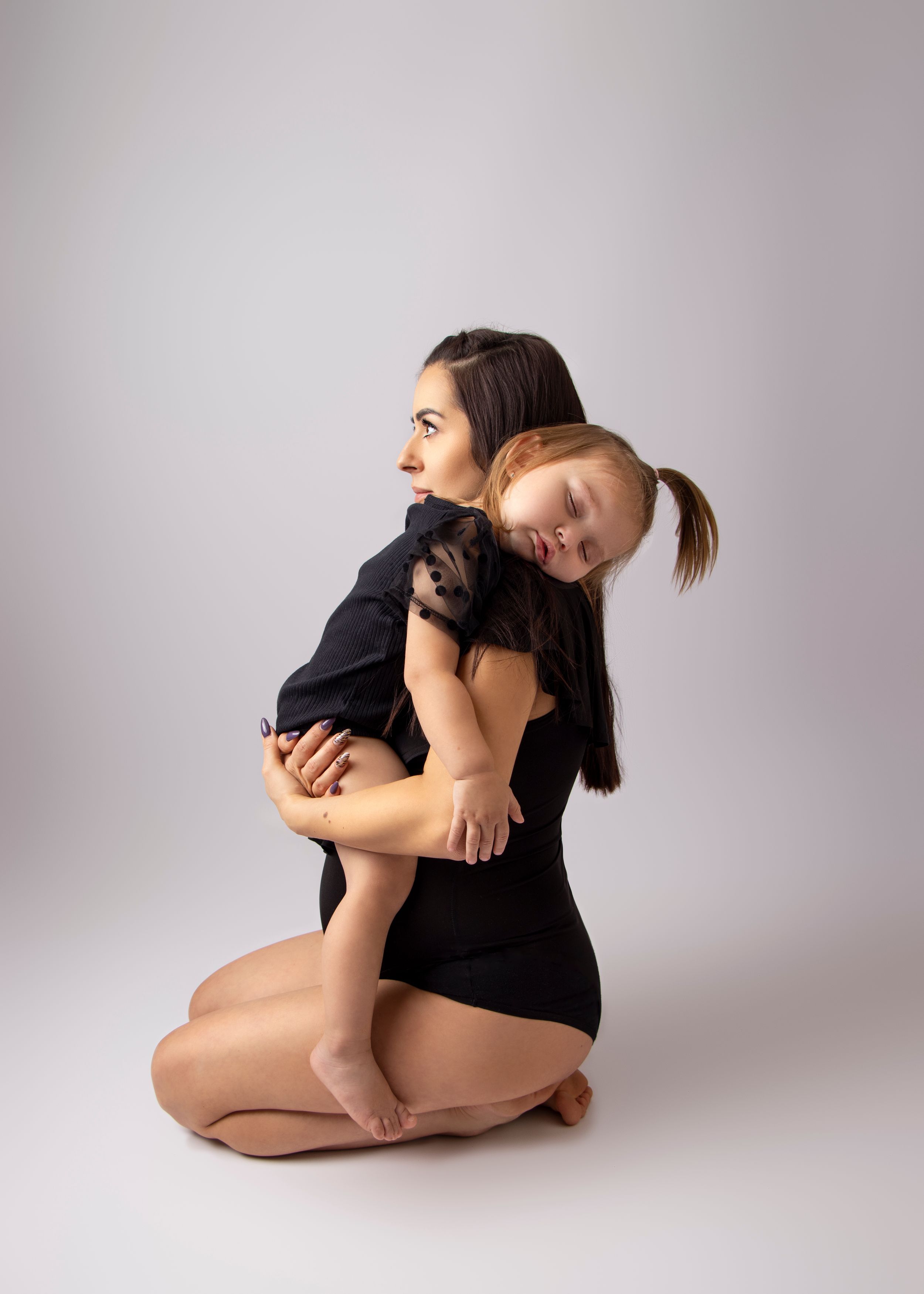 Mother and child share an intimate moment in black bodysuits against a gray backdrop.