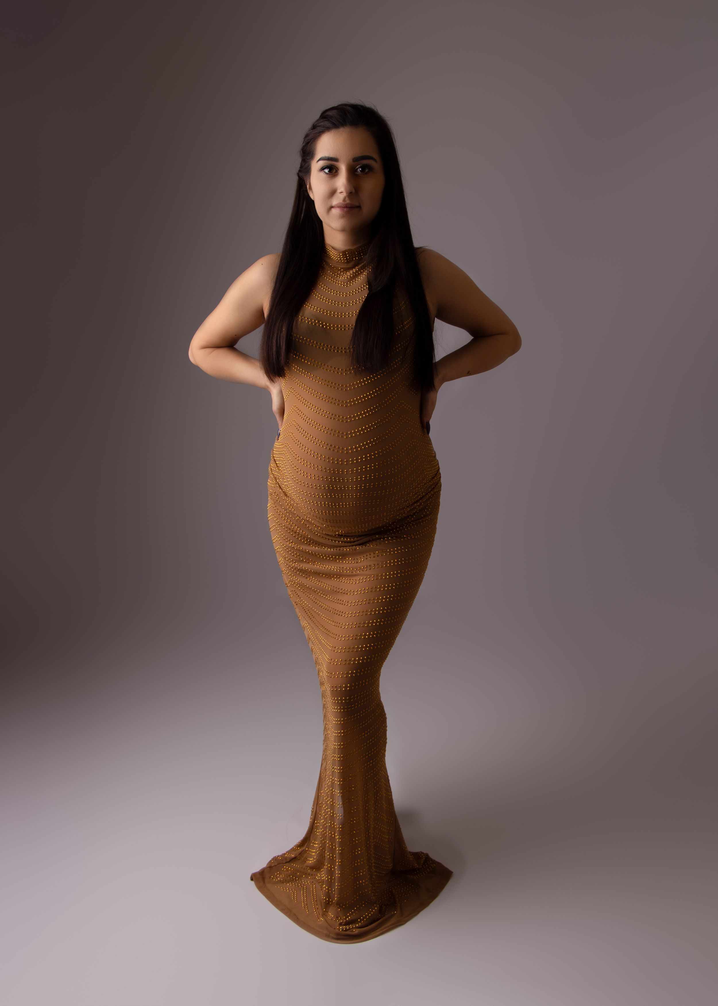 A full-length maternity portrait in a flowing brown dress against a neutral background.