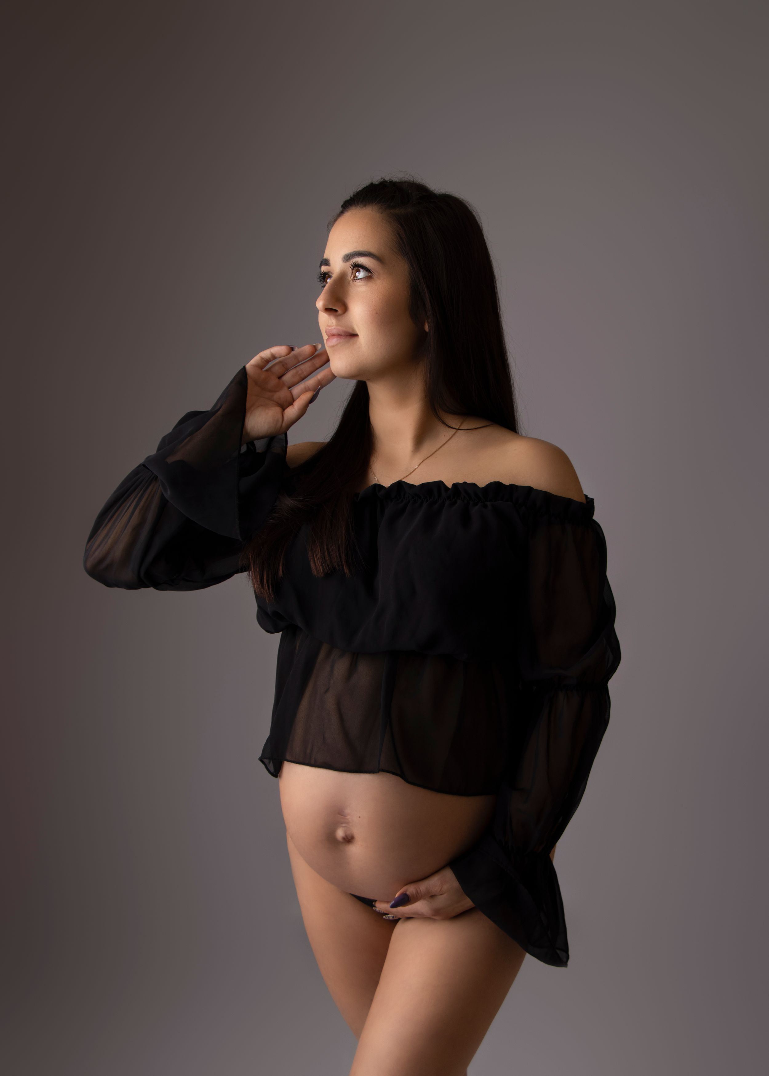 An artistic maternity portrait featuring a sheer black top against a neutral backdrop.