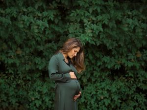 Pregnant woman in green dress stands against lush foliage background.