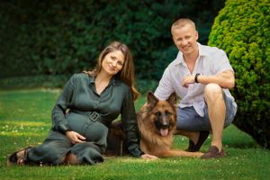 Expecting couple sits on grass with their German Shepherd dog in outdoor setting.