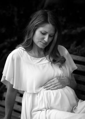 Expectant mother in white dress touches her belly while sitting on bench.
