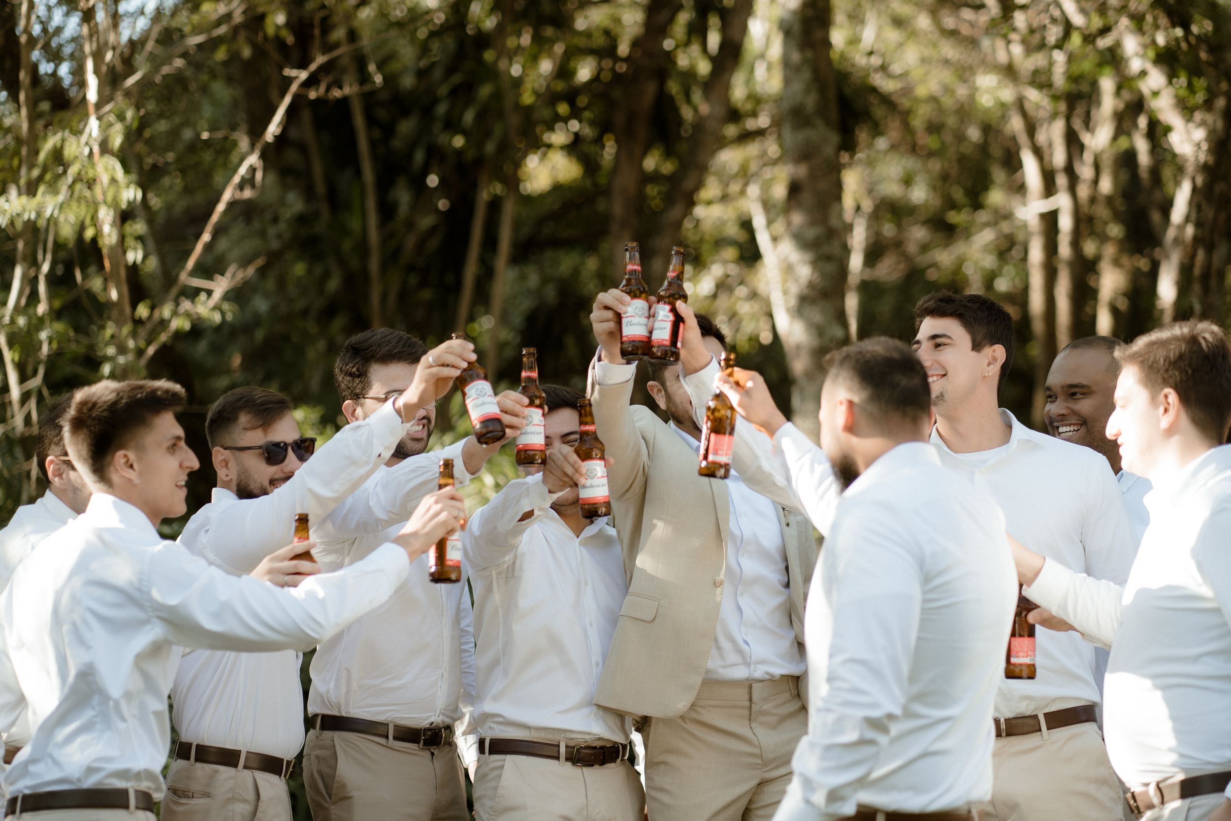 Group of groomsmen in white shirts and khaki pants toasting with drinks outdoors.