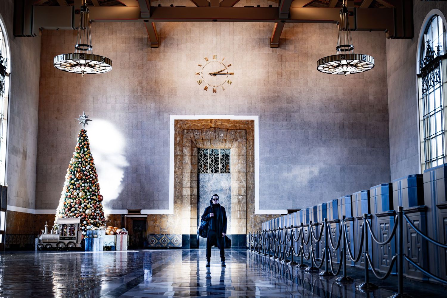 A silhouetted figure stands in a grand hall with a Christmas tree and arched windows.