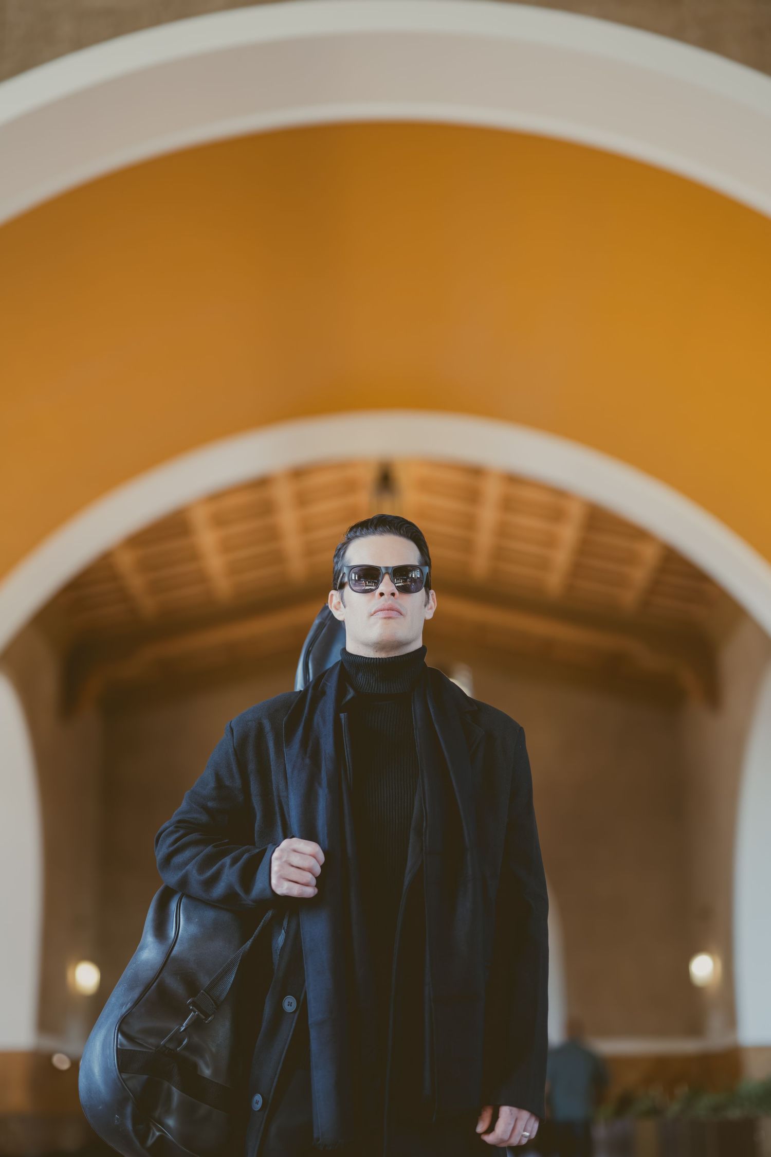 A person in a dark coat and sunglasses stands under an ornate archway carrying a bag.