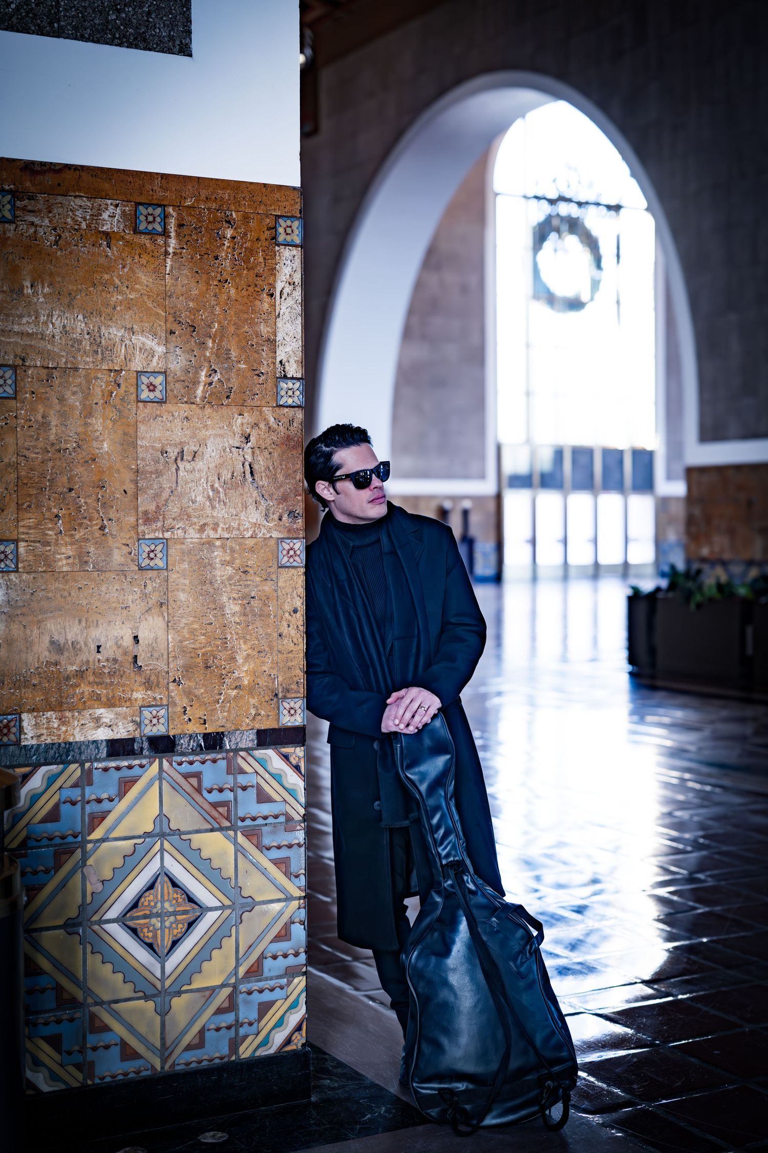 A person in dark clothing poses against decorative tiles under an arched walkway.