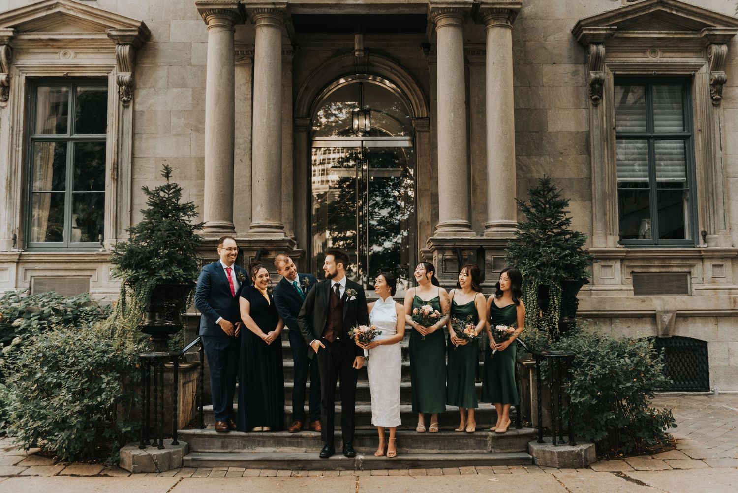 Wedding party stands on elegant stone steps of historic building with classical columns and evergreen shrubs.